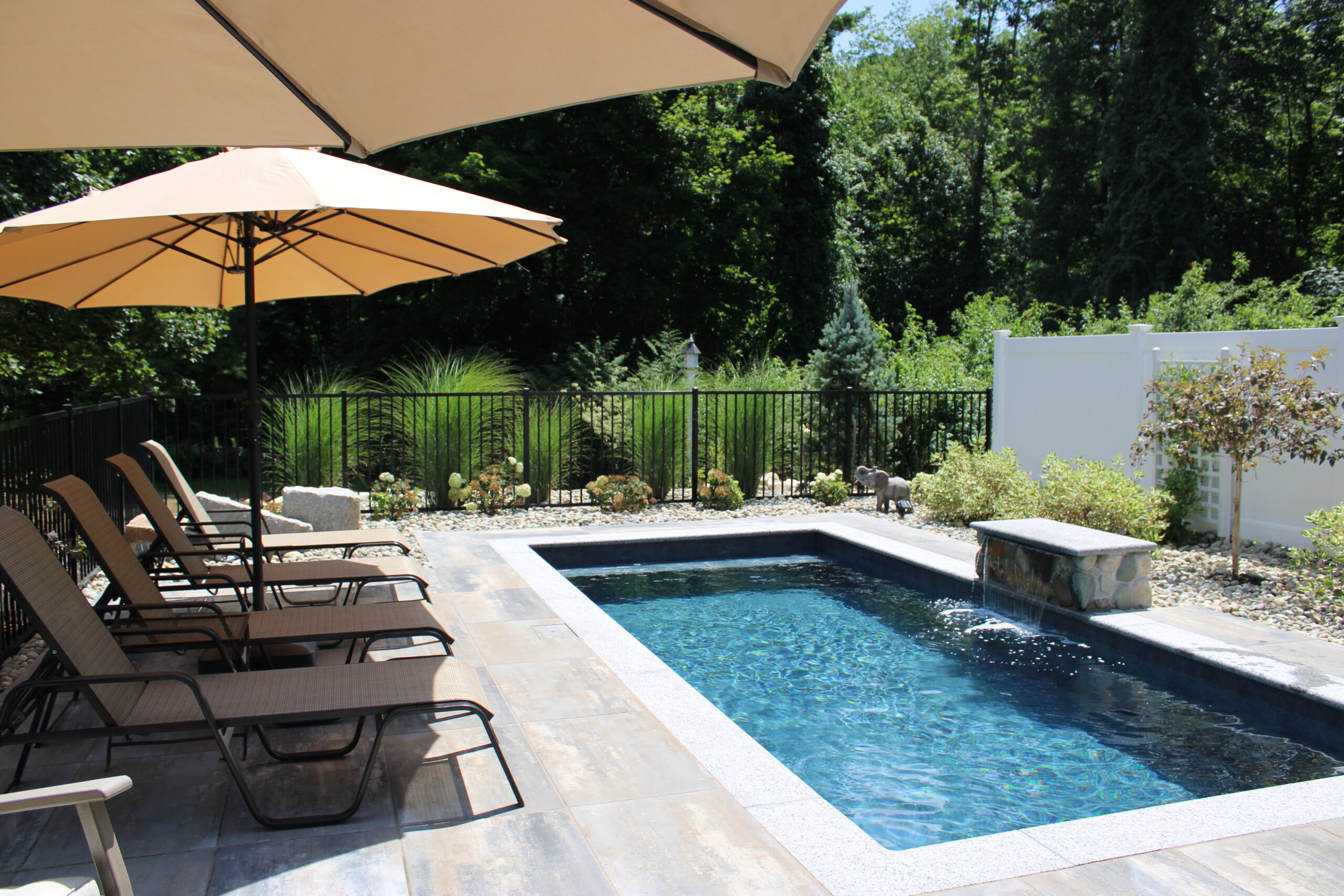 Modern plunge pool with RENOLIT interior finish, stone waterfall feature, and surrounding lounge chairs, umbrellas, and lush landscaping