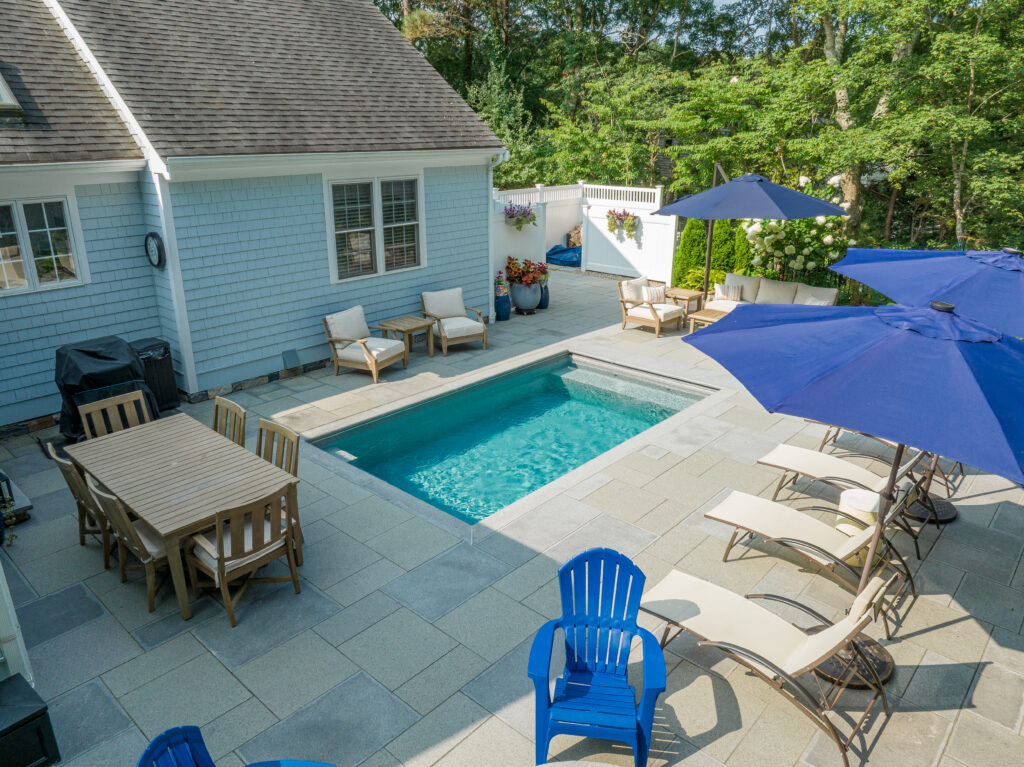 Backyard plunge pool with clear blue water, patio lounge chairs under navy umbrellas, outdoor dining table, and cushioned seating against a light blue shingle house.