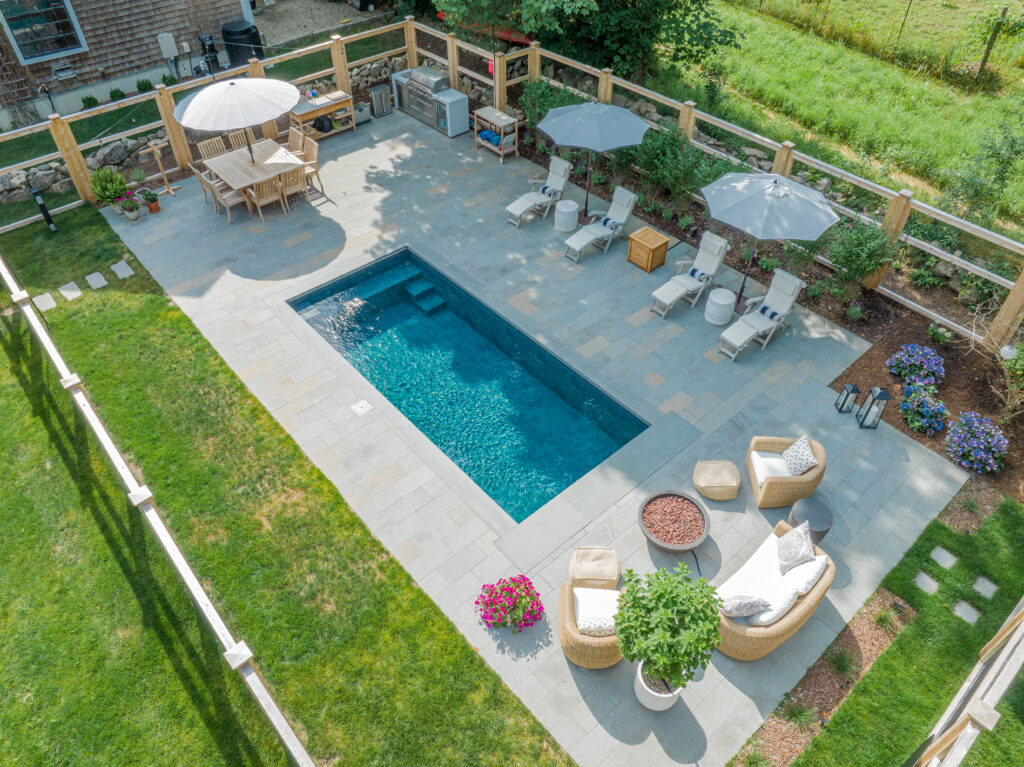 Aerial view of the Cape 9x21 plunge pool finished in Renolit, shade Elegance, featuring sparkling blue water, sleek stone patio, lounge chairs, umbrellas, and landscaped garden accents.