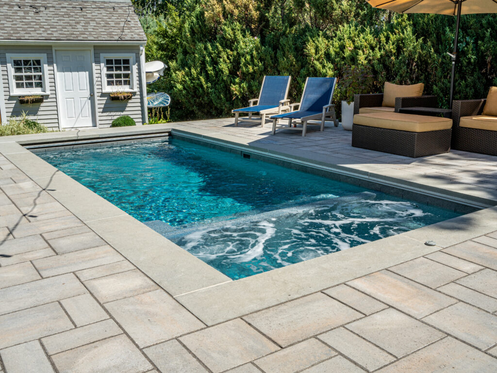 Rectangular backyard plunge pool with integrated spa, surrounded by light stone pavers. The poolside features lounge chairs, cushioned wicker seating, and a large umbrella, with a shed and greenery in the background.